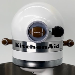May include: A white KitchenAid stand mixer with a football-shaped knob on the side and a football-shaped design on the front. The mixer is sitting on a stainless steel bowl.