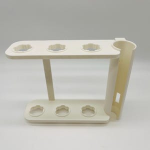 May include: Off-white plastic stand with a tiered design. The top tier has four circular openings with a scalloped edge, and the bottom tier has three similar openings. A vertical support with a curved section is on the right.