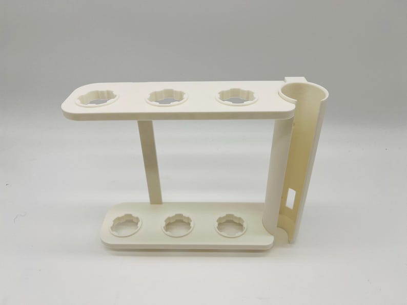 May include: Off-white plastic stand with a tiered design. The top tier has four circular openings with a scalloped edge, and the bottom tier has three similar openings. A vertical support with a curved section is on the right.