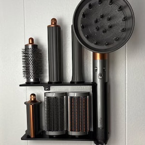 May include: A black and silver hair styling tool with multiple attachments, including a round brush, a comb, and a diffuser. The tool is mounted on a black wall-mounted stand. The brand name "Dyson" is visible on the tool.
