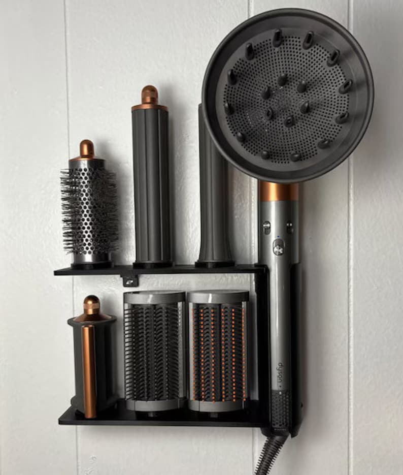 May include: A black and silver hair styling tool with multiple attachments, including a round brush, a comb, and a diffuser. The tool is mounted on a black wall-mounted stand. The brand name "Dyson" is visible on the tool.