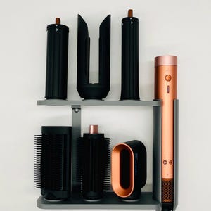 May include: A hair styling set featuring a copper and black hair dryer and various black styling attachments. The set is organized on a gray shelf against a white wall. The hair dryer has a copper-colored barrel.