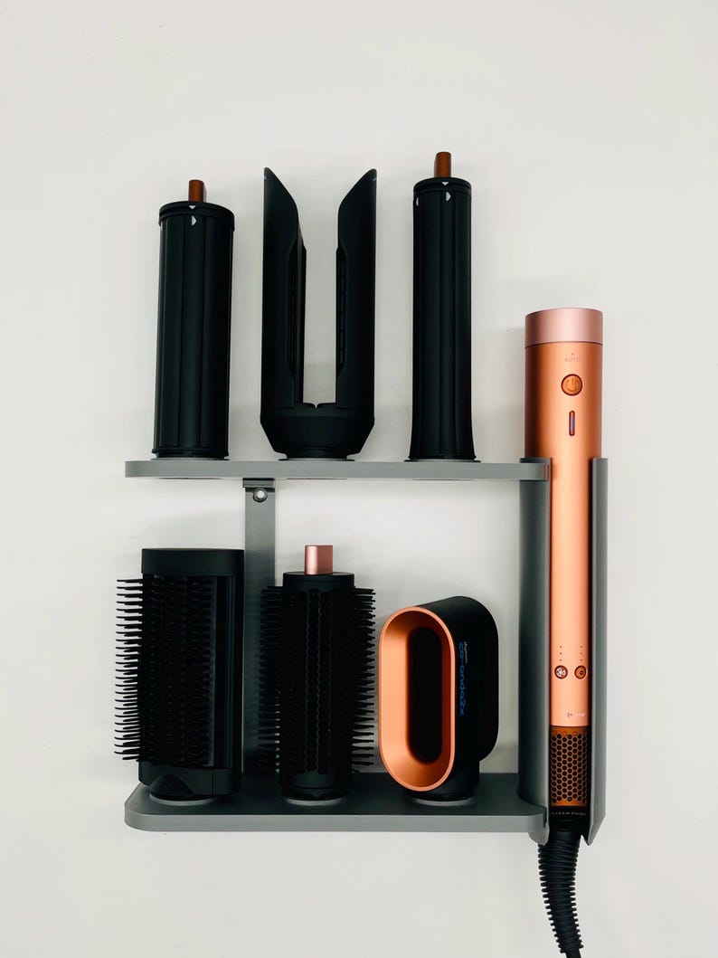 May include: A hair styling set featuring a copper and black hair dryer and various black styling attachments. The set is organized on a gray shelf against a white wall. The hair dryer has a copper-colored barrel.