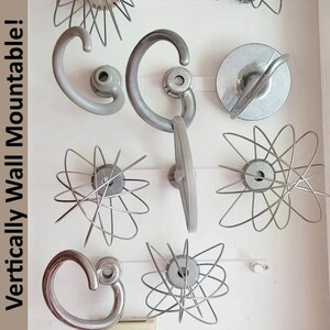 Op de afbeelding: Een verzameling vintage keuken-gardes en wandgrepen in verschillende ontwerpen. De gardes hebben draad lussen die uitstralen vanuit een centrale naaf. De handgrepen zijn gebogen en grijs. De tekst "Vertically Wall Mountable!" is zichtbaar links.