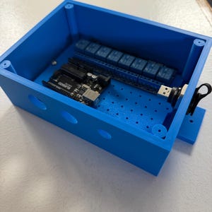 May include: A bright blue, rectangular electronic enclosure with a removable lid. Inside, an Arduino board and several blue relay modules are visible. The enclosure has several circular openings on the side and a USB port.