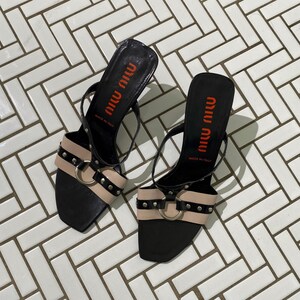 May include: A pair of black and beige heeled sandals with a silver buckle. The sandals have the text "Miu Miu" written on the straps in red. The text "Made in Italy" is also written on the straps.