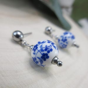 Peut inclure: Boucles d'oreilles pendantes avec des perles en céramique blanche ornées d'un motif floral bleu. Les boucles d'oreilles ont des tiges et des accents en métal argenté. Un design classique et délicat.