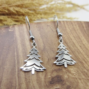 May include: A pair of silver-coloured Christmas tree earrings. The earrings feature a detailed cutout design of a fir tree, hanging from a simple hook. The earrings are displayed on a wooden surface.