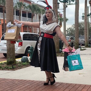 May include: A woman dressed in a black and white polka dot dress with a red belt and a white top. She is wearing a black feather hat and holding a shopping bag that says "House of Paws".
