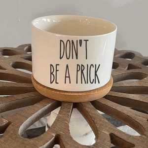 Puede incluir: Taza de cerámica blanca con un posavasos de bambú. La taza tiene un texto negro que dice "DON'T BE A PRICK".