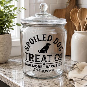 Könnte beinhalten: Ein durchsichtiges Leckerliglas mit Deckel, verziert mit einer schwarzen Grafik eines Hundes und dem Text "SPOILED DOG TREAT CO", "WAG MORE BARK LESS" sowie "50¢ a piece". Der zylindrische Behälter hat einen runden Glasknauf als Griff.