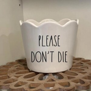 May include: White ceramic planter with a scalloped rim. The words "PLEASE DON'T DIE" are printed in black, block letters on the front. The planter sits on a decorative wooden stand.