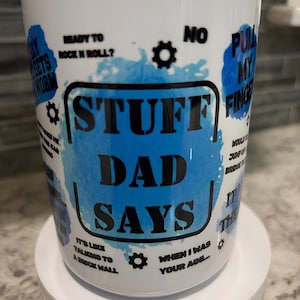 May include: White ceramic mug with the words "STUFF DAD SAYS" in black block letters, surrounded by blue watercolor-style accents and various phrases. The mug also features gear icons.
