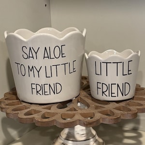 Puede incluir: Dos macetas de cerámica blanca con bordes festoneados. La maceta más grande dice "Say Aloe to My Little Friend" y la maceta más pequeña dice "Little Friend". Las macetas están sobre un pedestal de madera.