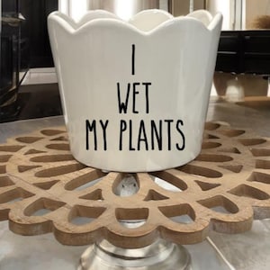 May include: A white ceramic planter with a scalloped rim, featuring the text "I WET MY PLANTS" in black. The planter sits on a decorative wooden stand with a floral design and a silver-colored base. The overall aesthetic is modern and suitable for indoor plants.