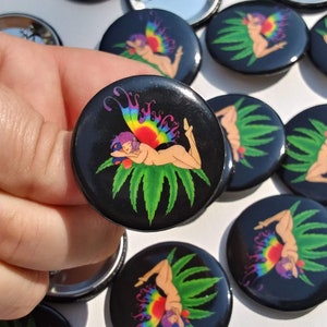 Puede incluir: Un conjunto de botones redondos negros y blancos con un hada colorida con alas de arcoíris tumbada sobre una hoja verde de marihuana. El hada lleva una corona de flores y tiene una expresión pacífica.