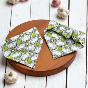 May include: Two triangular bandanas with a white garlic bulb pattern on a light green background, placed on a round wooden tray. The bandanas are surrounded by whole garlic bulbs on a white wooden surface.