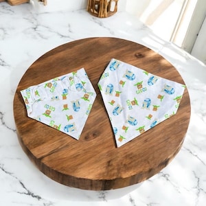 May include: Two white pet bandanas with a repeating pattern of Grogu, R2-D2, and other Star Wars characters in blue and green. The word "GROGU" is printed on the bandanas. The bandanas are on a wooden surface.