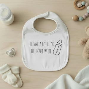 Puede incluir: Babero blanco para bebé con el texto "I'll take a bottle of the house white" y una ilustración de biberón. Rodeado de artículos para bebés, incluyendo un biberón, un chupete, calcetines y un osito de peluche.