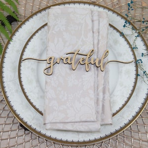 Puede incluir: Una tarjeta de lugar de madera con la palabra "grateful" escrita en cursiva. La tarjeta de lugar est&aacute; sobre una servilleta blanca en un plato blanco con un borde dorado.
