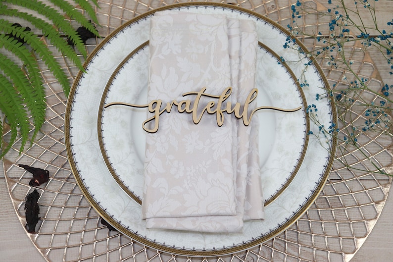 Puede incluir: Una tarjeta de lugar de madera con la palabra "grateful" escrita en cursiva. La tarjeta de lugar est&aacute; sobre una servilleta blanca en un plato blanco con un borde dorado.