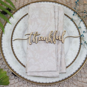 Puede incluir: Un marcador de lugar de madera con la palabra "thankful" escrita en cursiva. El marcador de lugar est&aacute; sobre una servilleta blanca en un plato blanco y dorado.
