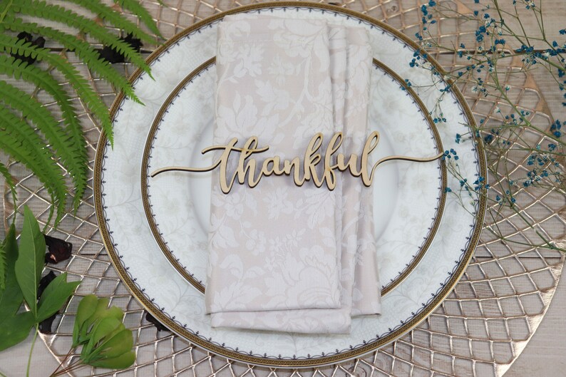 Puede incluir: Un marcador de lugar de madera con la palabra "thankful" escrita en cursiva. El marcador de lugar est&aacute; sobre una servilleta blanca en un plato blanco y dorado.