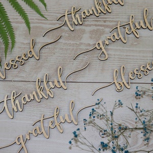 Puede incluir: Letreros de madera con las palabras "blessed", "thankful", "grateful" en una fuente cursiva. Los letreros est&aacute;n dispuestos sobre un fondo de madera clara con flores secas.
