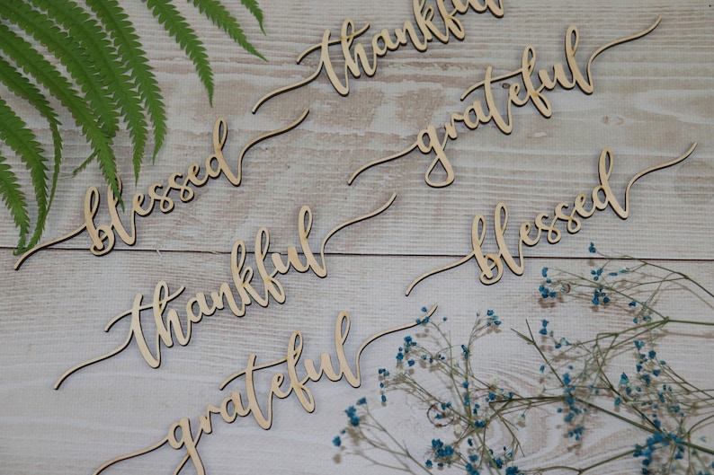 Puede incluir: Letreros de madera con las palabras "blessed", "thankful", "grateful" en una fuente cursiva. Los letreros est&aacute;n dispuestos sobre un fondo de madera clara con flores secas.