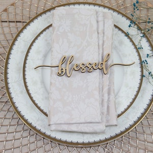 Puede incluir: Una tarjeta de lugar de madera con la palabra "blessed" escrita en cursiva. La tarjeta de lugar est&aacute; sobre una servilleta blanca en un plato blanco con borde dorado. El plato est&aacute; sobre un plato de base dorado.