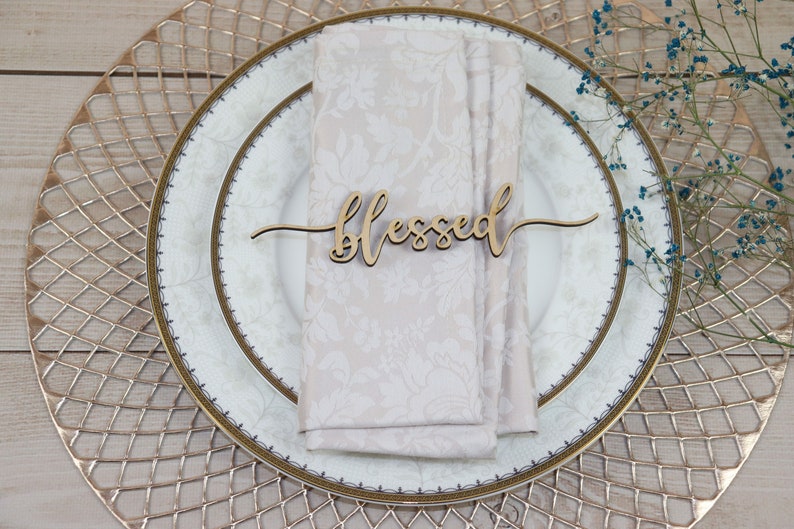 Puede incluir: Una tarjeta de lugar de madera con la palabra "blessed" escrita en cursiva. La tarjeta de lugar est&aacute; sobre una servilleta blanca en un plato blanco con borde dorado. El plato est&aacute; sobre un plato de base dorado.