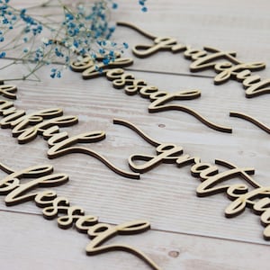 Puede incluir: Palabras recortadas de madera con las palabras "thankful", "blessed", "grateful" y "thankful" en una fuente cursiva. Las palabras est&aacute;n dispuestas en un patr&oacute;n escalonado sobre una superficie de madera clara.