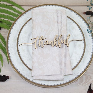 Puede incluir: Una servilleta blanca con ribete dorado se coloca sobre un plato blanco con ribete dorado. Un letrero de madera con la palabra "thankful" se coloca encima de la servilleta.