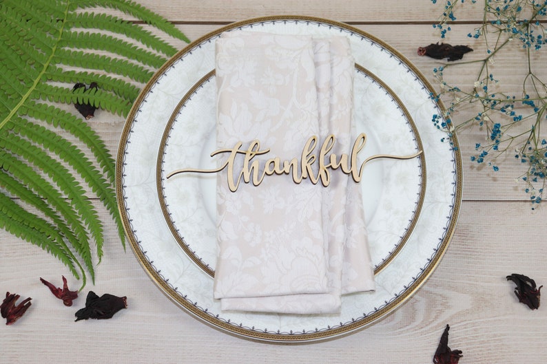 Puede incluir: Una servilleta blanca con ribete dorado se coloca sobre un plato blanco con ribete dorado. Un letrero de madera con la palabra "thankful" se coloca encima de la servilleta.