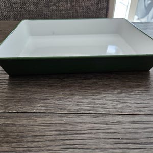 May include: A square baking dish with a white interior and a dark green exterior. The dish is made of metal and has a slightly sloped design. The dish is empty and sits on a wooden surface.
