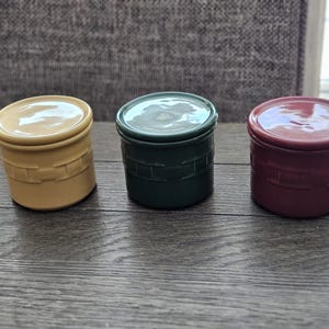 May include: Three small, lidded ceramic jars in mustard yellow, dark green, and burgundy. Each jar has a cylindrical shape with a textured, brick-like pattern around the base. The lids are smooth and rounded, creating a simple, rustic aesthetic.