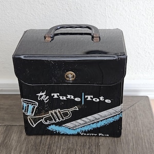 May include: Black vintage "Tune Tote" vanity case with a clear handle and a gold-colored clasp. The front features a blue and white design of a drum, trumpet, and piano keys. The text "the Tune | Tote" and "VANITY FAIR" are also visible.