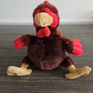 May include: A brown and red plush chicken toy with a red comb and wattle. The chicken is sitting on a wooden floor.