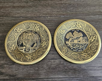 May include: Two round, gold-colored metal plates with intricate black designs. One plate features a black and white image of a building with Hebrew text. The other plate features a black and white image of a tree and a house with Hebrew text.