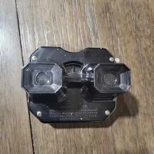 May include: A black View-Master viewer with two eye pieces and a central focus knob. The viewer has the text "Sawyer's View-Master" and other text on the front. The viewer is on a wooden surface.