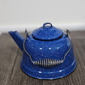 May include: A blue enamelware kettle with white speckles. The kettle has a spout, a lid with a small handle, and a wire handle with a coiled grip. The kettle is sitting on a wooden surface.
