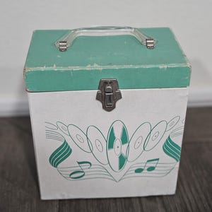 May include: Vintage record storage box with a teal green lid and a white body. The box features a clear plastic handle and a metal clasp. The front of the box is decorated with a green design of records and musical notes.