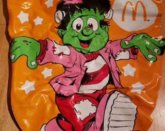 May include: Orange and white Halloween trick-or-treat bag with a green monster wearing a pink shirt and red shoes. The bag has the McDonald's logo on it.