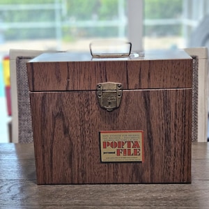 May include: A wooden file box with a handle and a metal clasp. The box has a wood grain pattern and a label that reads "Porta File". The label also states "Safe Storage for Receipts, Tax Records, Documents".