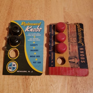May include: Two vintage packages of replacement knobs for cabinets, drawers, bread boxes, and pot lids. The packages are both red, white, and blue with black text. The left package shows three black knobs and the right package shows four red knobs.
