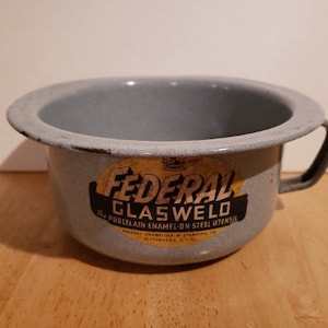May include: A gray enamelware bowl with a black handle. The bowl has a gold sticker that reads "Federal Glasweld The Porcelain Enamel-on-Steel Utensil Federal Enameling & Stamping Co. Pittsburgh, Pa. U.S.A."