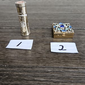 May include: A gold-colored cylindrical container with text and a red top, next to a small, square, gold-colored box adorned with blue and clear gemstones. Two white paper notes with the numbers 1 and 2 are also present.