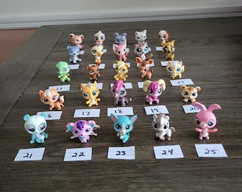 Puede incluir: Una colección de 25 mascotas de juguete de plástico de colores, cada una con un diseño único y numerada en una tarjeta blanca. Los juguetes están dispuestos en filas sobre una superficie de madera.