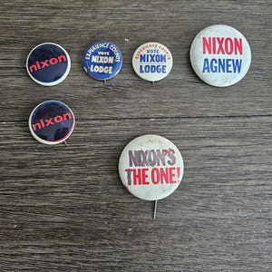 May include: Six vintage political campaign buttons featuring the names "Nixon" and "Agnew". The buttons are white with red and blue lettering. One button reads "Nixon's The One!"