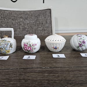May include: Four white ceramic decorative items on a wooden surface. Includes a small teapot with a handle, a jar with a lid, a small bowl with a lid, and a round item. The teapot has a gold accent and a map design. The jar and round item have floral designs.